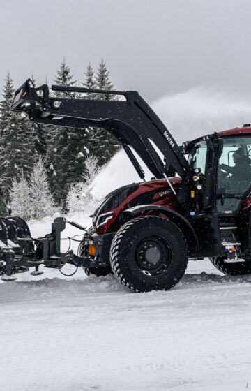 Snøplog montert på traktor rydder snø på gårdsplass i vinterlandskap.